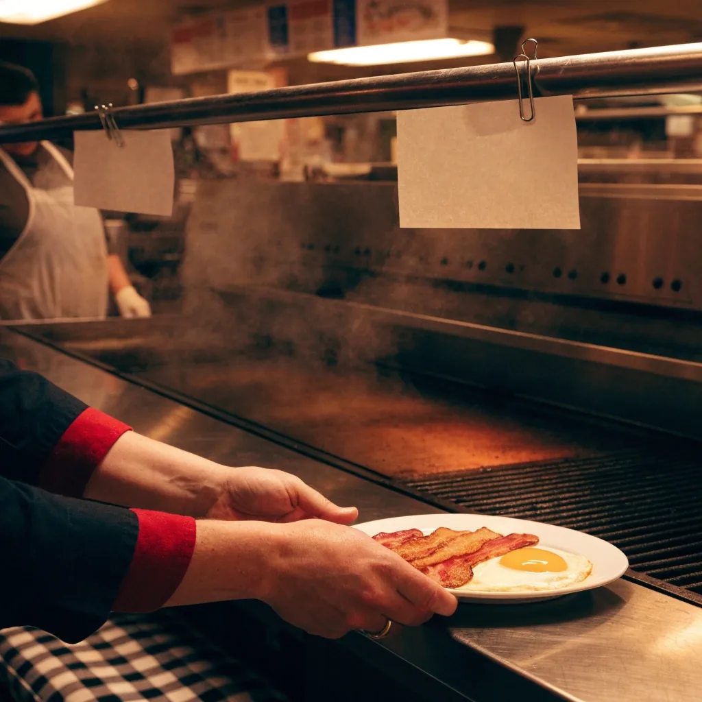 A cook's hands sliding a finished plate forward beneath blank order slips clipped on the rail